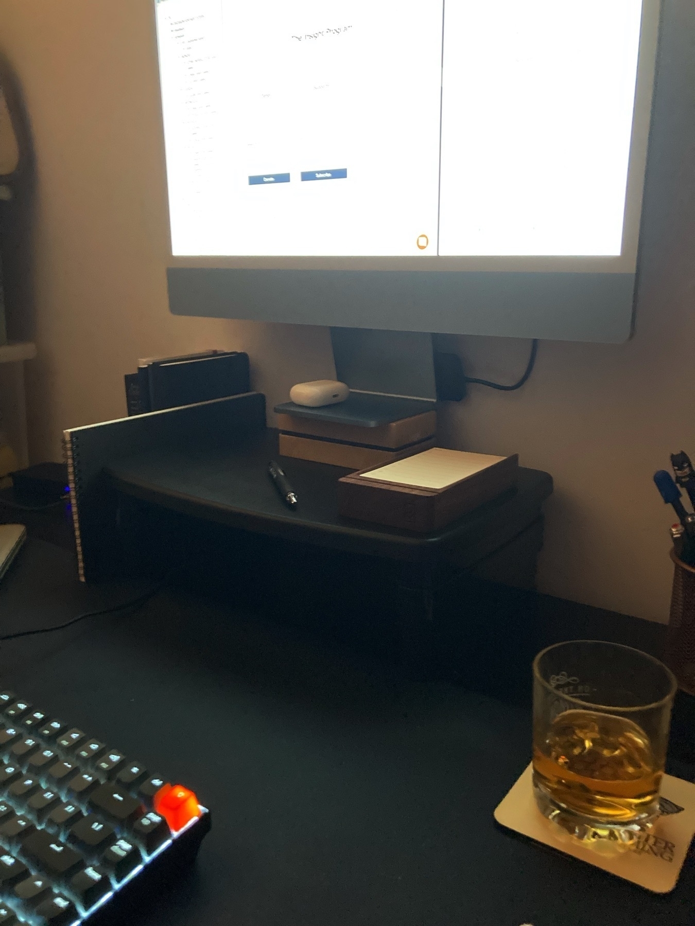 Part of a desk, including a computer, keyboard, pen, AirPods case, glass of whiskey on a coaster, notebooks, and the Analog productivity system.