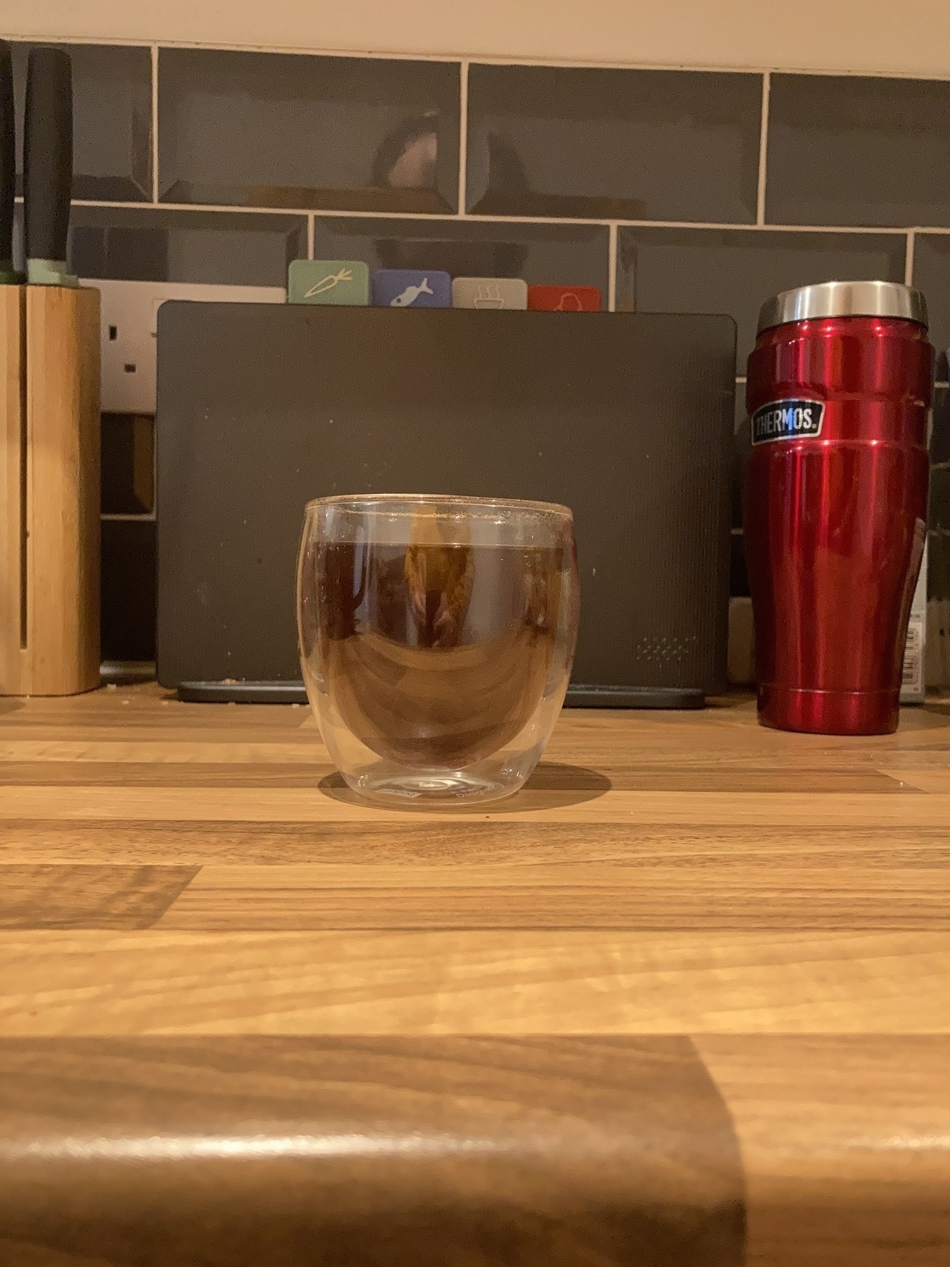 A glass cup of coffee atop a kitchen counter, with chopping boards, a metal thermal container, and a knife block in the background.
